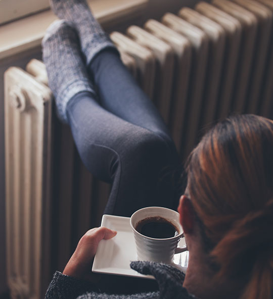 A close-up of a person sitting by a radiator with a cup of tea