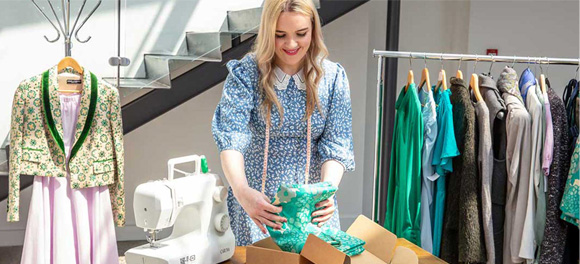 A female business owner in a blue dress packages a green dress for an eCommerce order.